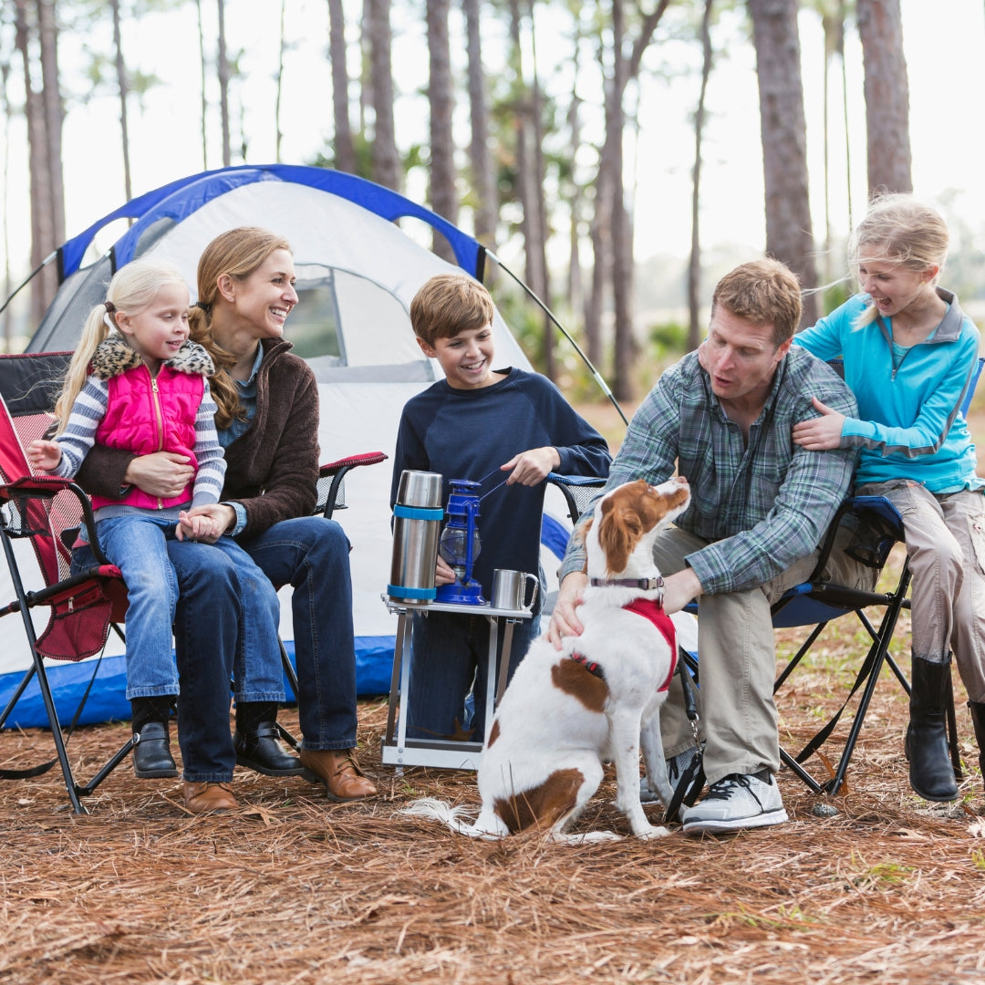 A family of four and their dog are camping outdoors in a pine forest next to a blue and white dome tent. The mother is holding a young girl, the father is petting a brown and white dog wearing a red bandana, and a boy stands by a small folding table with metal mugs.