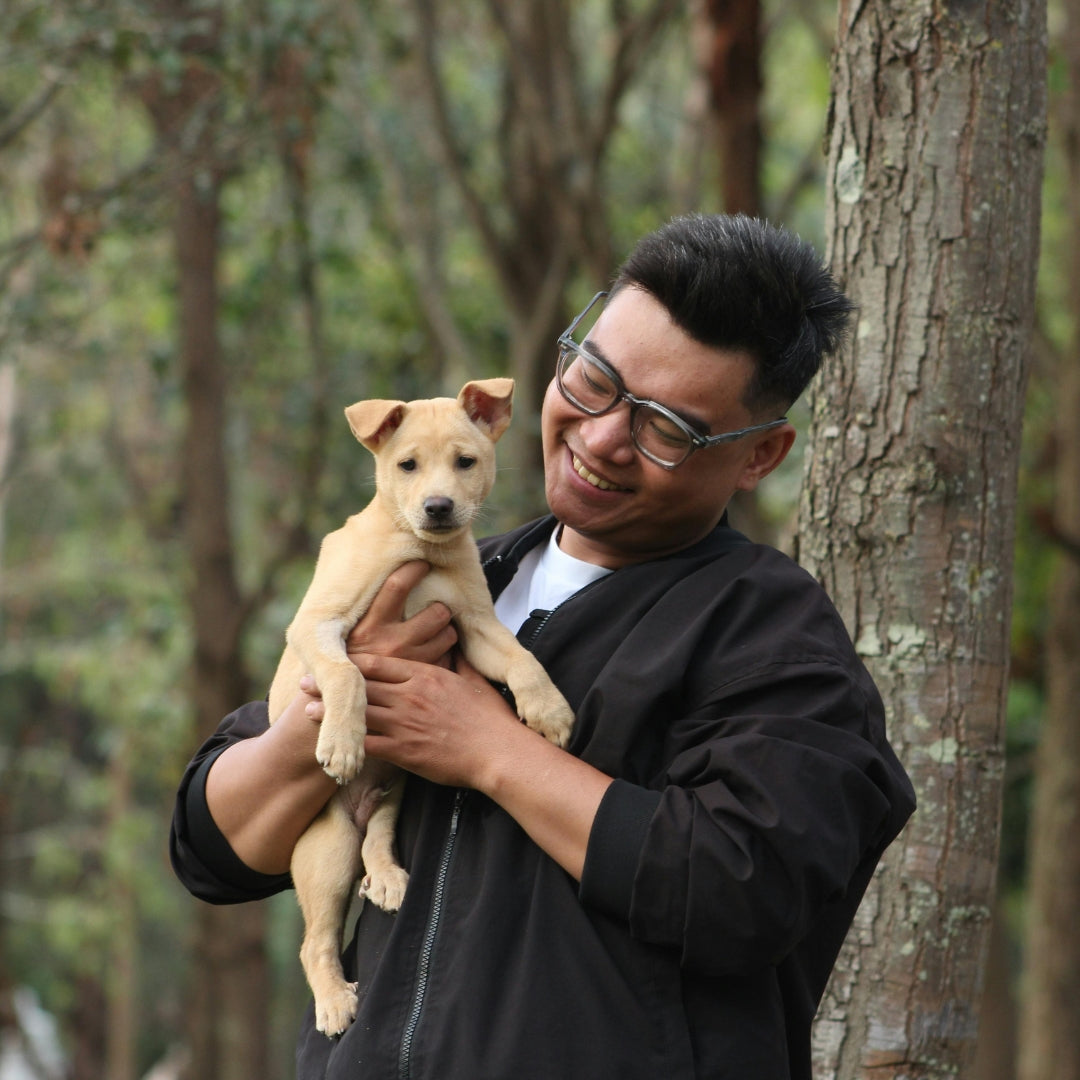 A man wearing glasses and a black jacket holds a small light tan puppy in his arms, standing outdoors in front of a tree trunk with a blurred forest background.