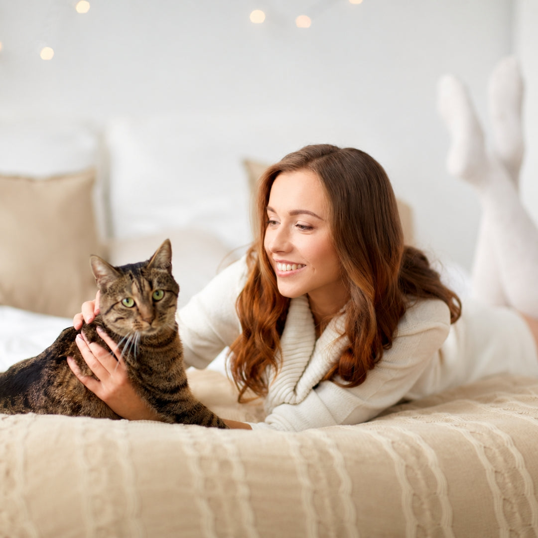 A smiling woman lies on a bed with a brown tabby cat, wearing a cream knit sweater and white socks in a softly lit bedroom.