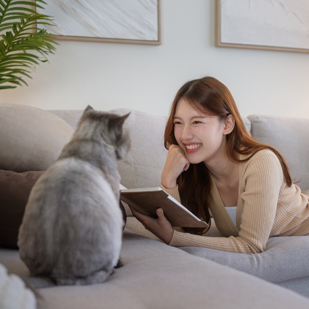 A woman smiling while lying on a light gray sofa and holding a tablet, with a gray cat sitting in the foreground facing her. The setting is a bright, modern living room with framed wall art and a plant visible.