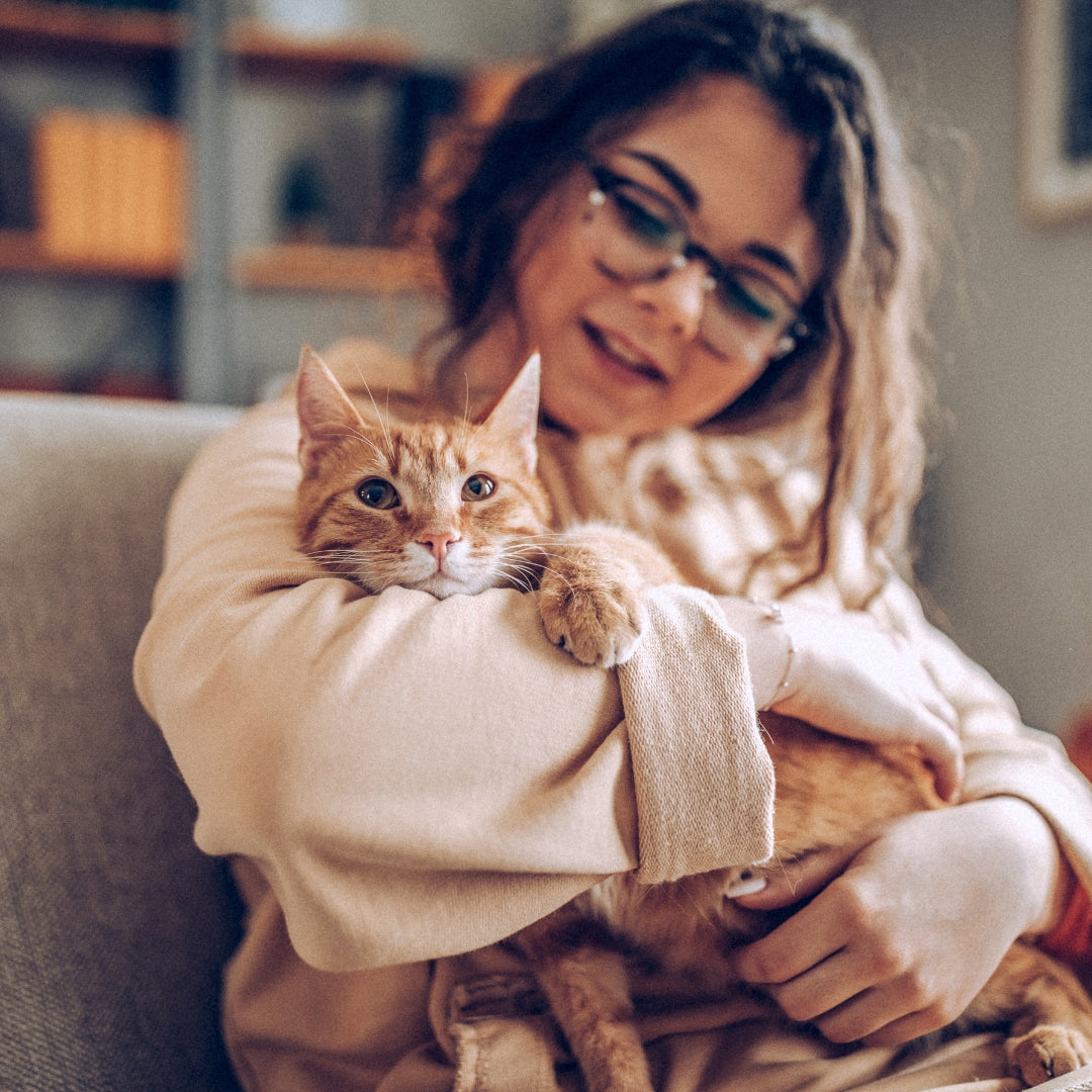 A woman wearing glasses and a beige knit sweater holds an orange tabby cat in her lap while sitting on a couch in a cozy indoor setting.