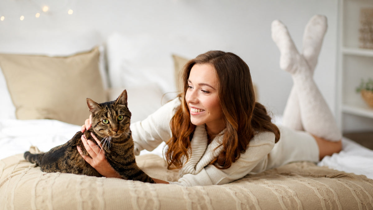 A woman in a cozy white sweater and white socks lies on a bed with a brown tabby cat, smiling warmly in a bright, softly lit bedroom.