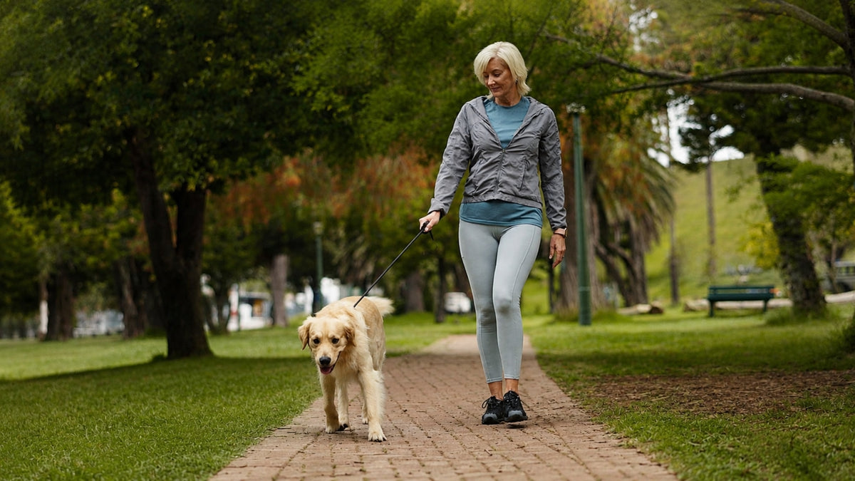 A woman walking a light-colored dog on a paved path in a park, wearing a gray zip-up hoodie, blue t-shirt, and light gray leggings.