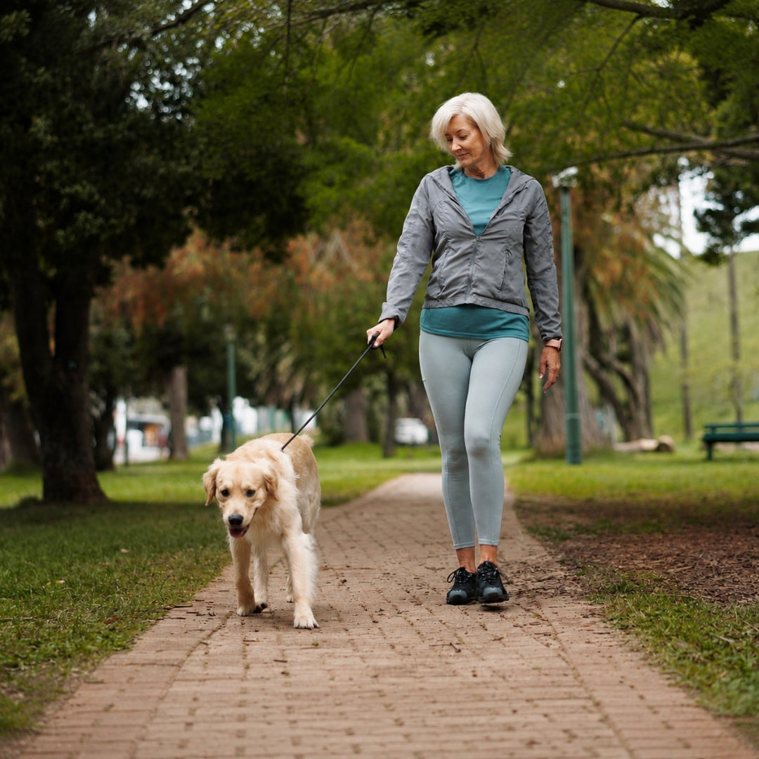 A woman walking a golden retriever on a brick path in a park, wearing a gray zip-up hoodie over a teal top and light gray leggings.
